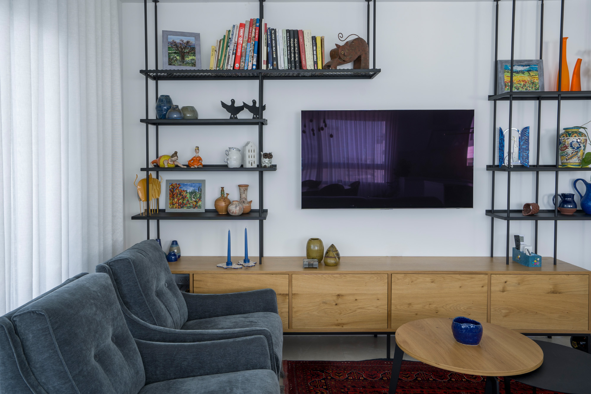 חדר מגורים מודרני ומסודר עם יחידת טלוויזיה עשויה עץ, מדפי מתכת שחורים עם חפצים דקורטיביים, כורסאות בד כחולים ושולחן קפה עגול על שטיח אדום.