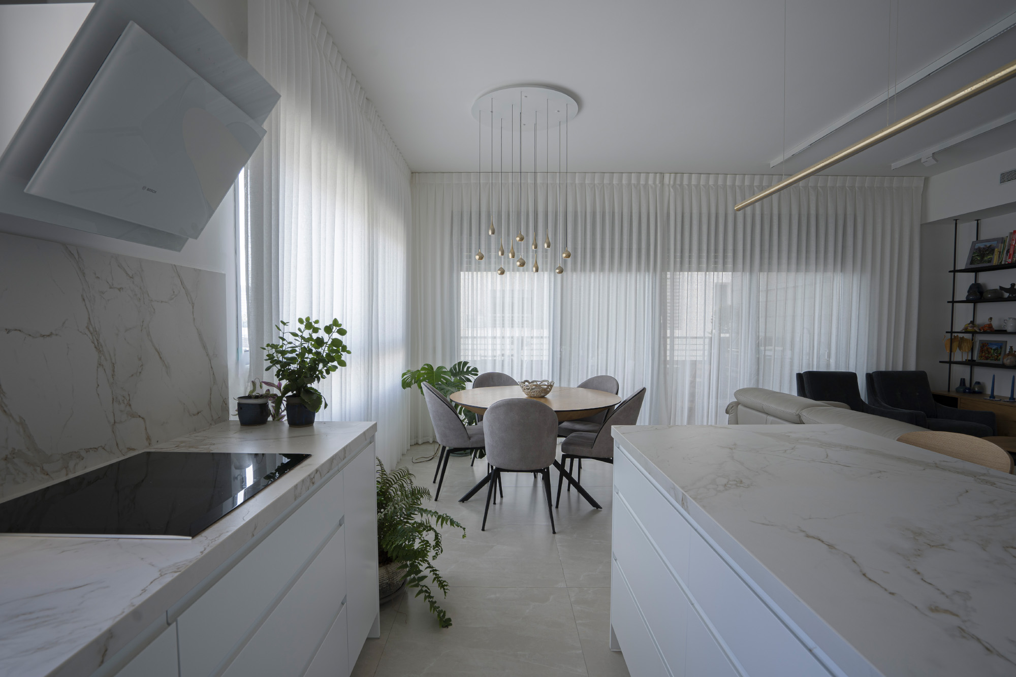 מטבח מודרני עם משטח שיש לבן, כיריים שחורים ומנדף, לצד פינת אוכל עם שולחן עגול וכיסאות אפורים, מוארים במנורת תלייה מעוצבת, וחלון גדול עם וילונות לבנים ברקע.