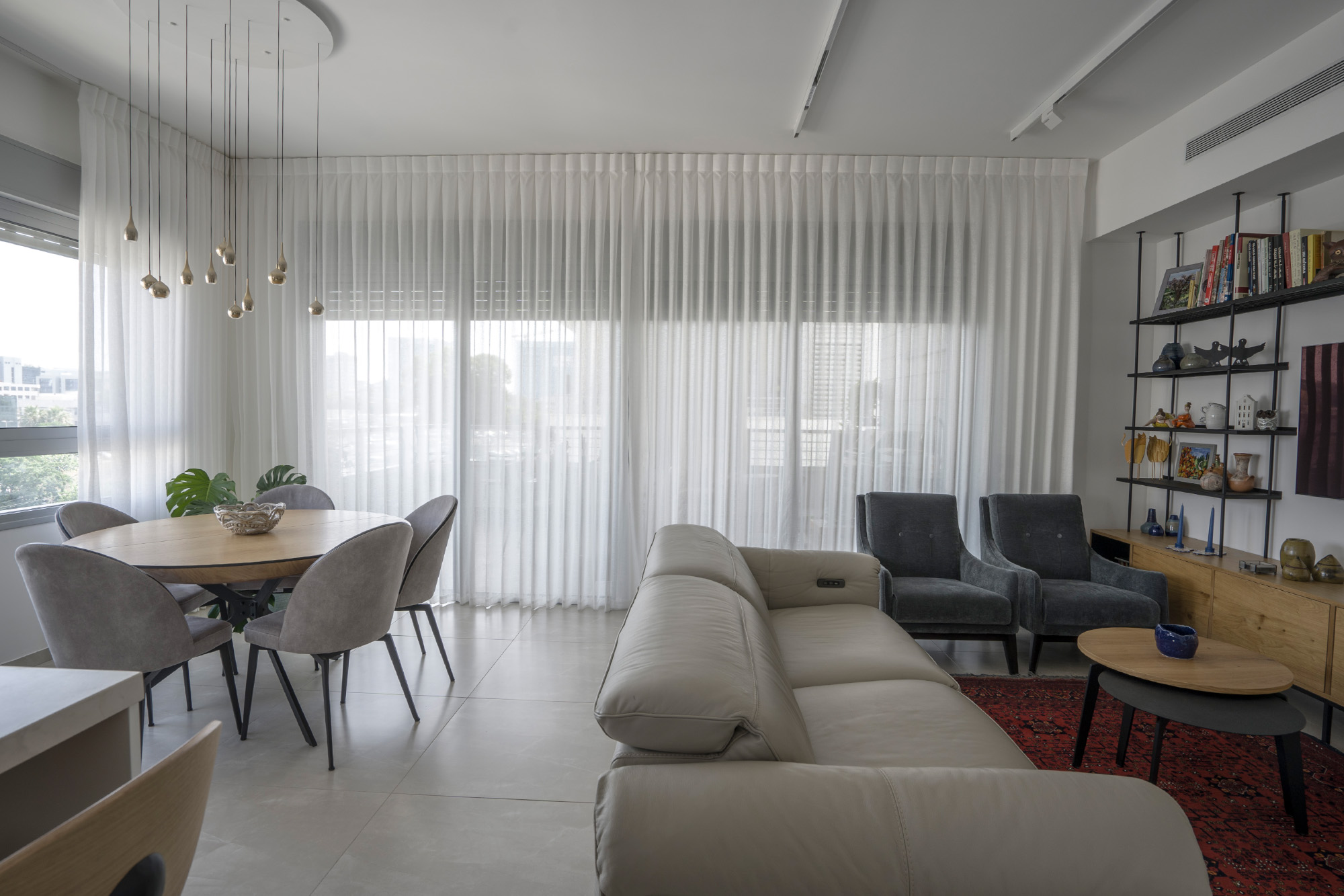 חלל מואר עם ספה אפורה, כורסאות כחולים, שולחן קפה ושולחן אוכל עגול עם כיסאות אפורים, מואר במנורת תלייה. וילונות לבנים, שטיח אדום ומדפים שחורים עם חפצים דקורטיביים מוסיפים סגנון.